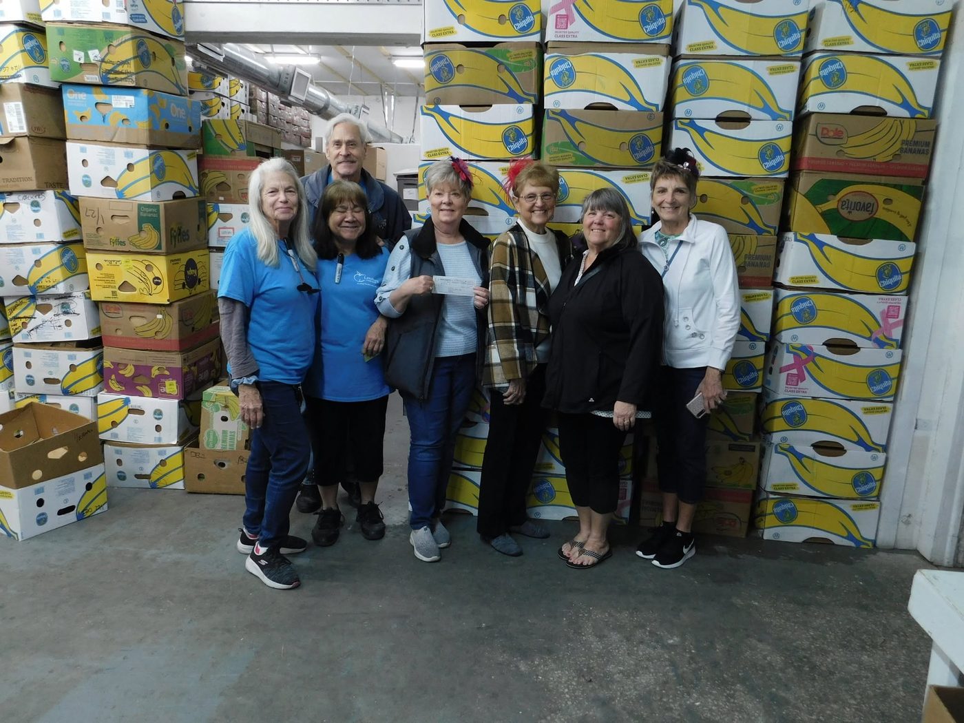 Leesburg Food Bank volunteers accepting a donation check in the warehouse, surrounded by stacked banana boxes used for food storage.
